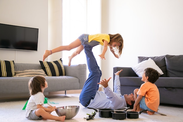 Pai feliz segurando a filha nas pernas e deitada no tapete. Alegres crianças brancas brincando na sala de estar com o pai. Dois meninos bonitos sentados no chão. Conceito de atividade de infância, férias e jogos