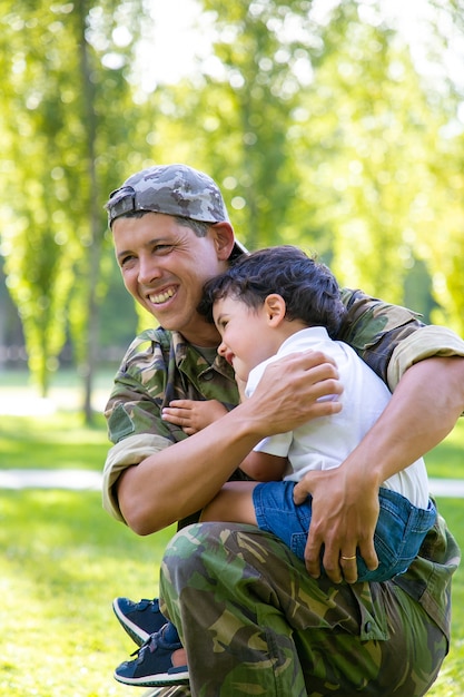 Pai militar alegre segurando o filho nos braços, abraçando o menino ao ar livre, após retornar da viagem missionária. Tiro vertical. Conceito de reunião familiar ou retorno a casa