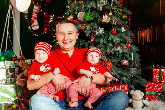 Pai senta com gêmeos em ternos vermelhos antes de uma árvore de Natal