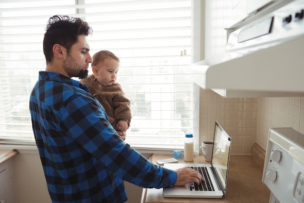 Foto grátis pai usando laptop enquanto segura seu bebê na cozinha