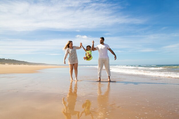 Pais alegres e uma menina curtindo caminhadas e atividades na praia, criança segurando as mãos dos pais, pulando e jogando as pernas para cima