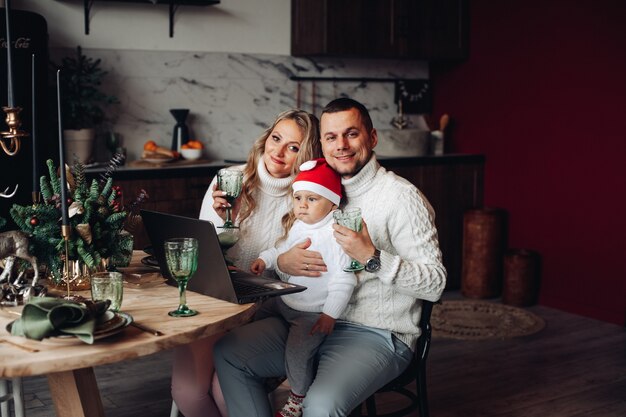 Pais e filho fofo com chapéu de Papai Noel segurando óculos enquanto jantam de Natal e usando laptop