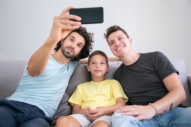 Foto grátis pais homossexuais alegres e criança tomando selfie na cela, sentado no sofá em casa, sorrindo para a câmera frontal. vista frontal. família e conceito de comunicação