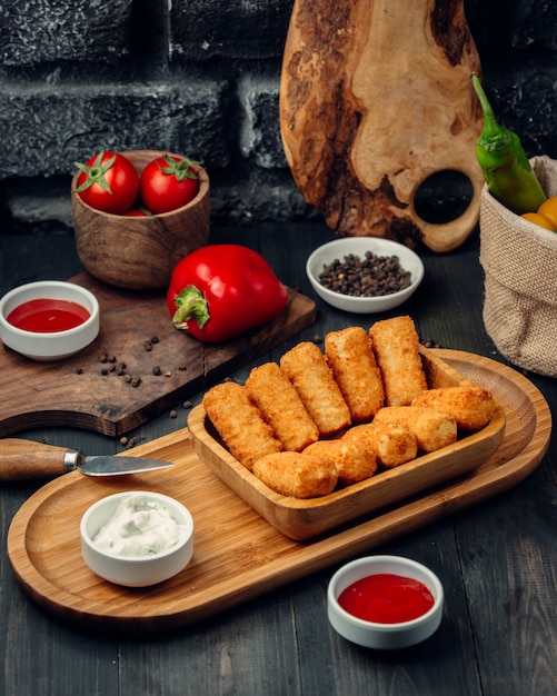 Palitos de frango frito em uma placa de madeira com maionese e molho de tomate.