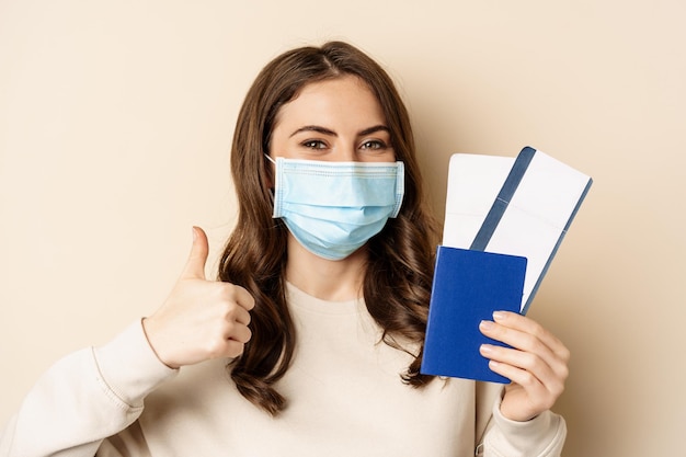 Pandemia de viagens e covid-19. Turista de mulher feliz com máscara médica, mostrando os polegares para cima e passaporte com dois ingressos, fundo bege.