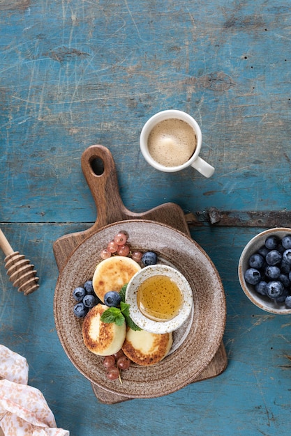 Panquecas de queijo cottage, cheesecakes, bolinhos de ricota com mirtilos frescos, groselhas e pêssegos em um prato Café da manhã saudável e delicioso para o feriado Fundo azul de madeira