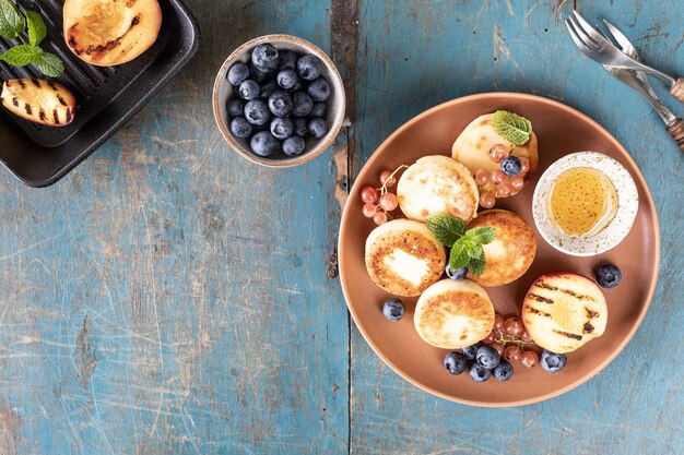 Panquecas de queijo cottage, cheesecakes, bolinhos de ricota com mirtilos frescos, groselhas e pêssegos em um prato Café da manhã saudável e delicioso para o feriado Fundo azul de madeira