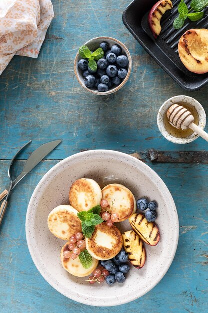 Panquecas de queijo cottage, cheesecakes, bolinhos de ricota com mirtilos frescos, groselhas e pêssegos em um prato Café da manhã saudável e delicioso para o feriado Fundo azul de madeira