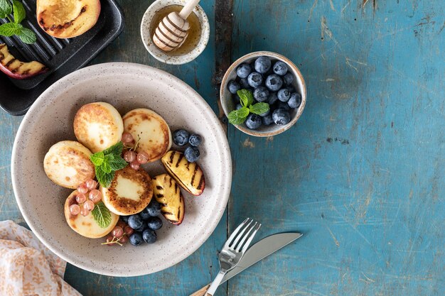 Panquecas de queijo cottage, cheesecakes, bolinhos de ricota com mirtilos frescos, groselhas e pêssegos em um prato Café da manhã saudável e delicioso para o feriado Fundo de madeira azul