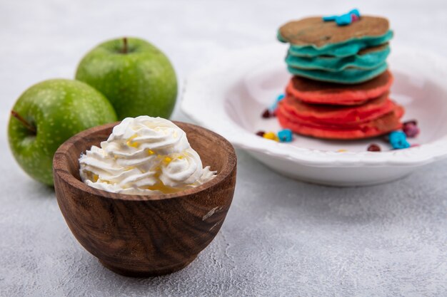 Panquecas multicoloridas de vista frontal em um suporte com maçãs verdes e chantilly em um pires em um fundo branco