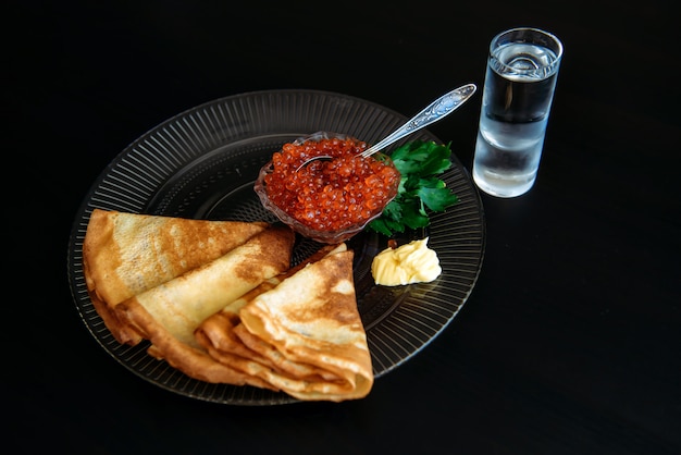Panquecas russas com caviar vermelho e manteiga no prato transparente perto de copo de vodka de gelo isolada no preto