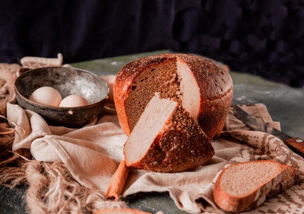Pão redondo e fatiado inteiro em uma mesa de cozinha de pedra com ovos e faca.