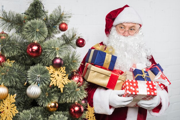 Papai Noel com caixas de presente nas mãos perto da árvore de Natal decorada
