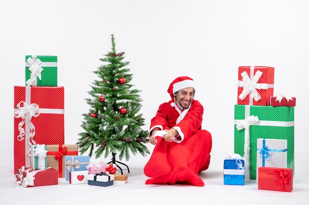 Papai Noel sentado no chão segurando uma meia de natal perto de presentes e uma árvore decorada