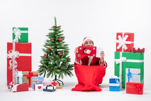 Papai Noel sorridente, sentado no chão e levantando a meia de natal na cara, perto de presentes e decorado com uma árvore de ano novo em fundo branco