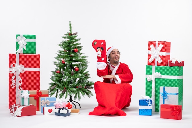 Papai Noel sorridente, sentado no chão, usando uma meia de natal perto de presentes e decorando a árvore de ano novo em fundo branco