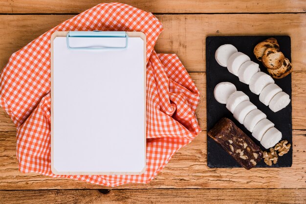 Papel em branco branco na prancheta com pano de mesa perto de queijo de cabra e lanche sobre pedra ardósia