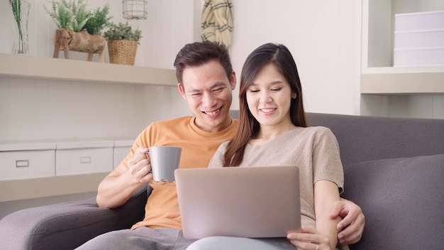 Par asiático, usando computador portátil, e, bebida, xícara quente café, em, sala de estar, em, lar, doce, par, desfrute