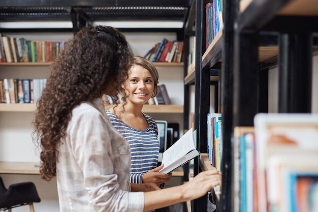 Par de meninas lindas em roupas elegantes casuais em pé perto de estantes de livros na biblioteca, olhando um ao outro, falando sobre a vida universitária tentando encontrar literatura para a lição de amanhã.