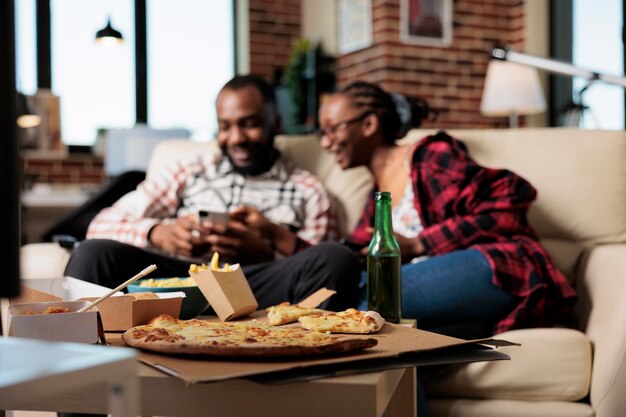 Parceiros afro-americanos com comida para viagem usando telefones celulares para navegar na internet, assistindo filmes na televisão e comendo entrega de fast food. Atividade de lazer com take away e tv.