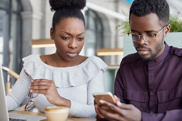 Parceiros de negócios masculinos e femininos de pele escura sérios verificar notificação no telefone inteligente, trabalhar no relatório no computador portátil, sentar no café têm olhares concentrados.