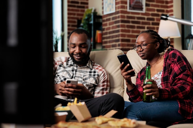 Parceiros millennials navegando na internet no celular e assistindo tv, comendo fast food para viagem. Pacote de refeições para comer e assistir filme na televisão juntos.