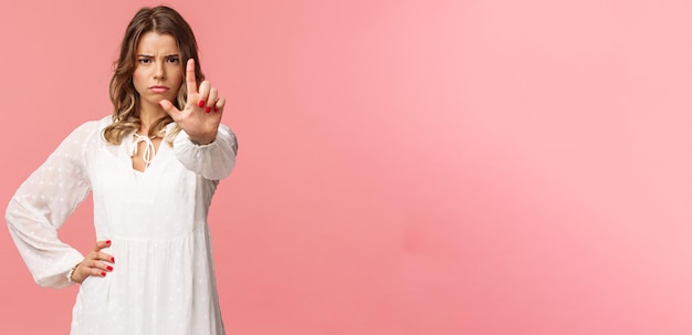 Foto grátis pare o suficiente retrato de uma garota loira assertiva e forte de aparência séria no vestido branco estende a mão em direção à câmera com um dedo levantado em advertência franzindo a testa com raiva proibir e proibir a ação