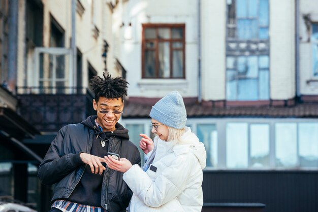 Pares adolescentes inter-raciais à moda que fazem o divertimento olhando no telefone móvel