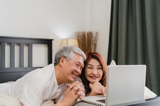 Pares sênior asiáticos usando o laptop em casa. As avós chinesas sênior asiáticas, o marido e a esposa felizes após acordam, vendo o filme deitado na cama no quarto em casa no conceito da manhã.