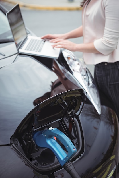 Parte média da mulher usando laptop enquanto carrega o carro elétrico