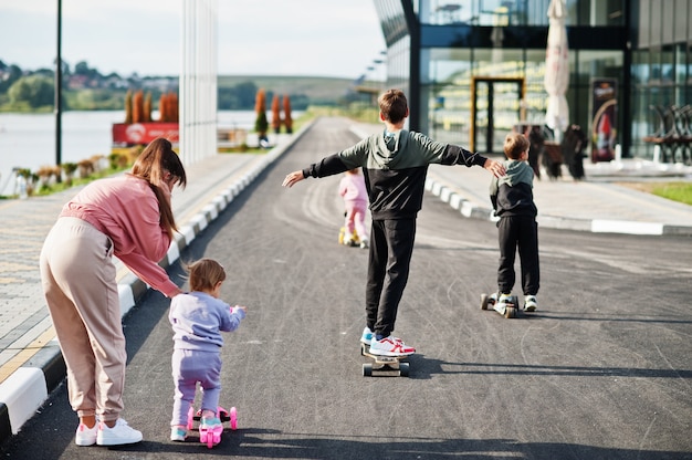 Parte traseira da jovem mãe elegante com quatro filhos ao ar livre. a família de esportes passa o tempo livre ao ar livre com patinetes e patins.