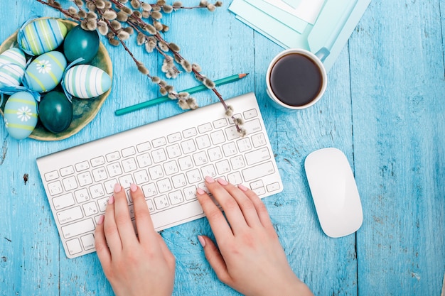 Páscoa no local de trabalho de escritório, mesa de madeira azul. mão feminina em um teclado de computador e uma xícara de café