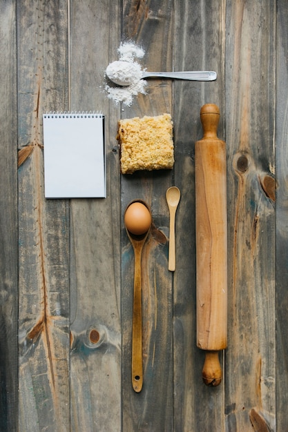 Pastelaria; rolo de massa; colher; ovo; farinha e bloco de notas na superfície de madeira