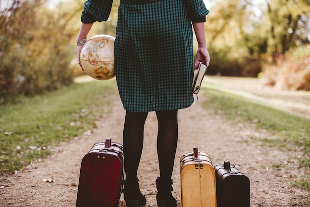 Pé feminino em uma estrada vazia perto de sua mala velha, segurando a bíblia e um globo de mesa