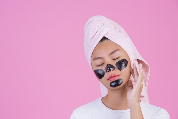 Peeling cosmético de beleza. A fêmea nova com preto retira a máscara no produto cosmético da casca dos cuidados com a pele da pele no rosto em um rosa.