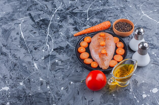 Peito de frango e cenouras fatiadas em um prato ao lado de sal, óleo, especiarias, cenoura e tomate na superfície azul