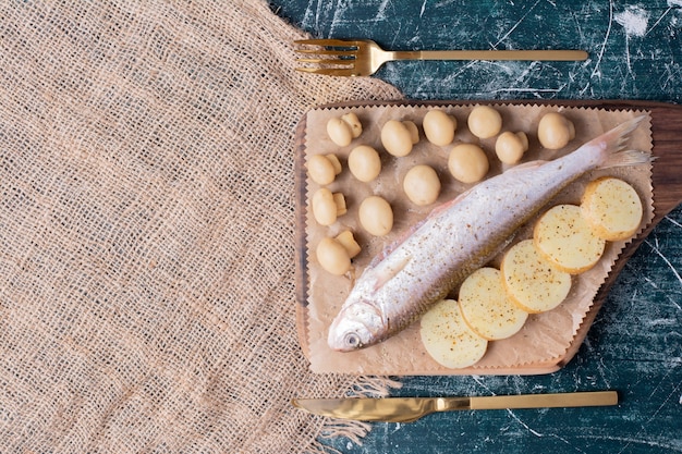 Peixes inteiros crus com azeitonas e rodelas de batata cozida na placa de madeira.