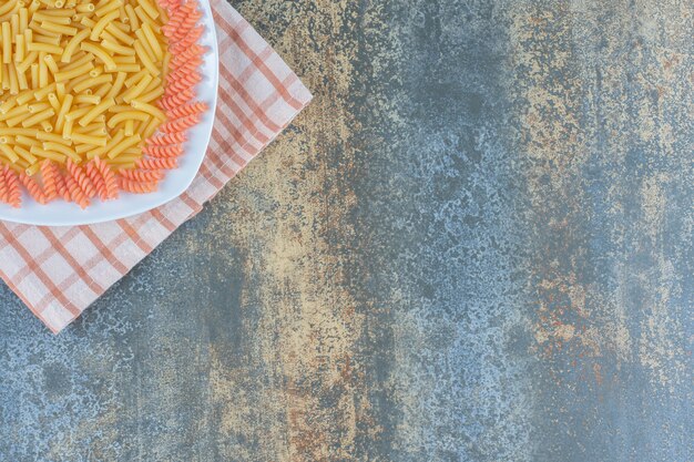 Penne e massa fusilli crus em tigela sobre toalha, sobre a superfície de mármore.