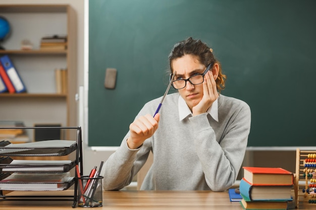 Pensando colocando a mão no queixo jovem professor masculino de óculos segurando o ponteiro sentado na mesa com ferramentas escolares em sala de aula