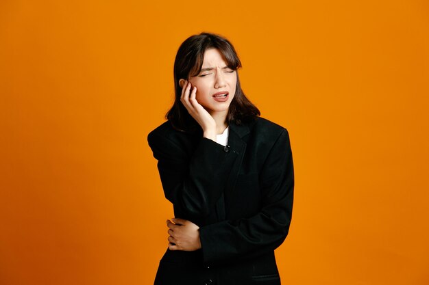 Pensando em colocar a mão no queixo, jovem linda mulher vestindo jaqueta preta isolada em fundo laranja