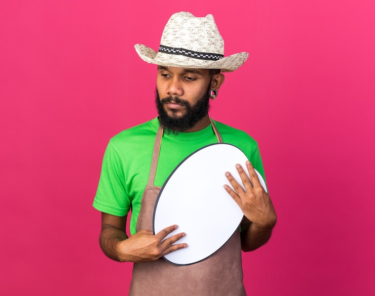 Foto grátis pensando em olhar ao lado, jovem jardineiro afro-americano, usando um chapéu de jardinagem, segurando um balão de fala isolado na parede rosa