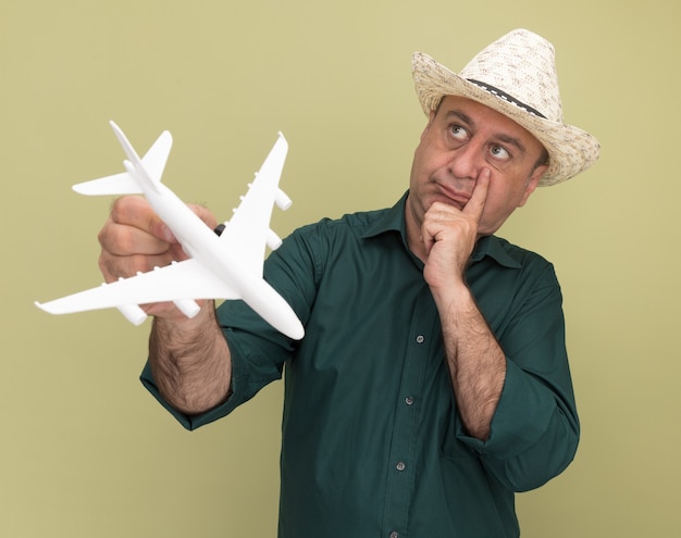 Pensando em olhar para o lado homem de meia-idade vestindo camiseta verde e chapéu segurando um avião de brinquedo colocando a mão na bochecha isolada na parede verde oliva