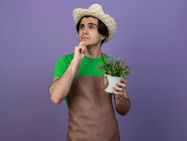 Foto grátis pensando em olhar para o lado, jovem jardineiro de uniforme, usando chapéu de jardinagem, segurando uma flor em um vaso de flores, agarrou o queixo