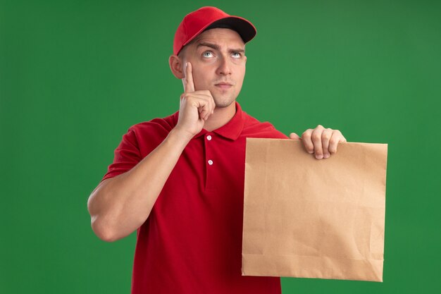 Pensando em olhar para um jovem entregador de uniforme e boné segurando um pacote de comida de papel, colocando o dedo na bochecha isolada na parede verde