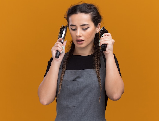 Pensando em olhar para uma jovem barbeira de uniforme segurando o pente com a tesoura de cabelo ao redor do rosto isolado na parede laranja