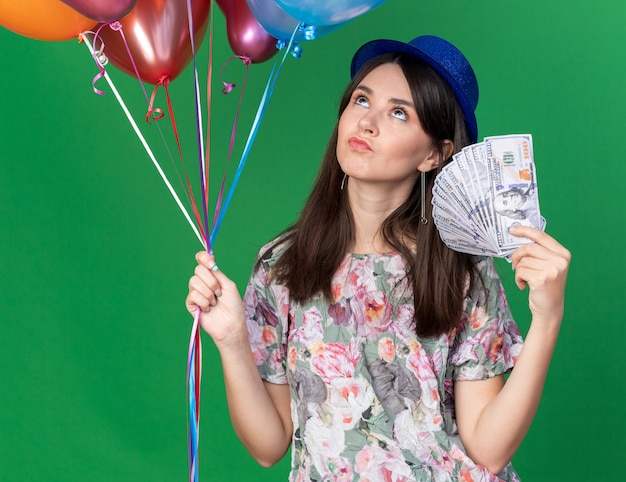 Pensando em olhar uma jovem linda com chapéu de festa segurando balões com dinheiro isolado na parede verde
