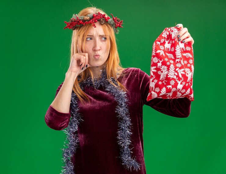 Foto grátis pensando em uma linda garota com um vestido vermelho com grinalda e guirlanda no pescoço segurando e olhando para a bolsa de natal colocando a mão na bochecha isolada no fundo verde