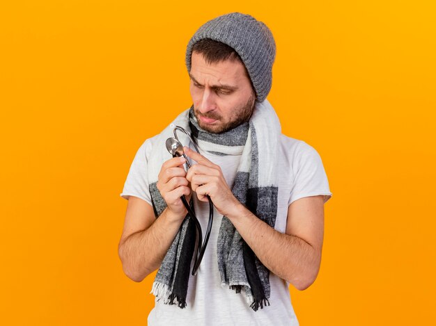 Pensando jovem doente com chapéu de inverno e lenço segurando e olhando para o estetoscópio isolado no amarelo