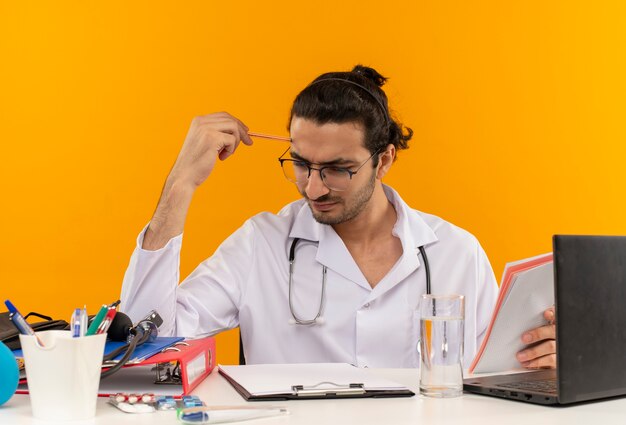 Pensando jovem médico com óculos médicos, usando roupão médico com estetoscópio sentado na mesa