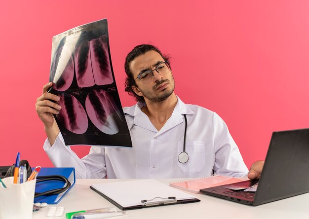 Pensando jovem médico com óculos médicos, usando roupão médico com estetoscópio sentado na mesa